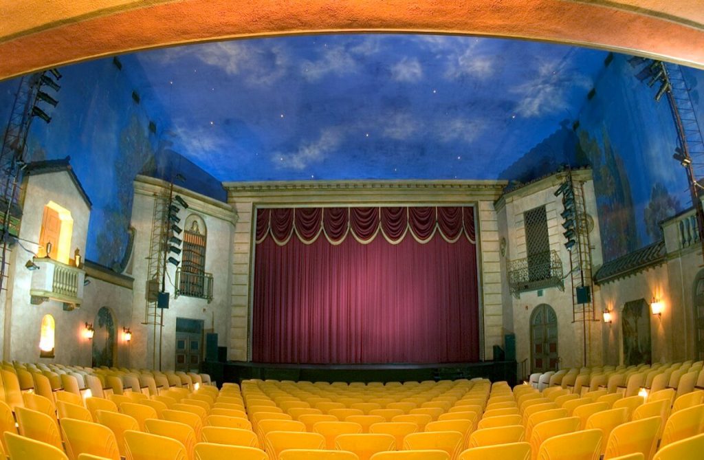 large theater with rows of yellow chairs and red stage curtain and blue ceiling.