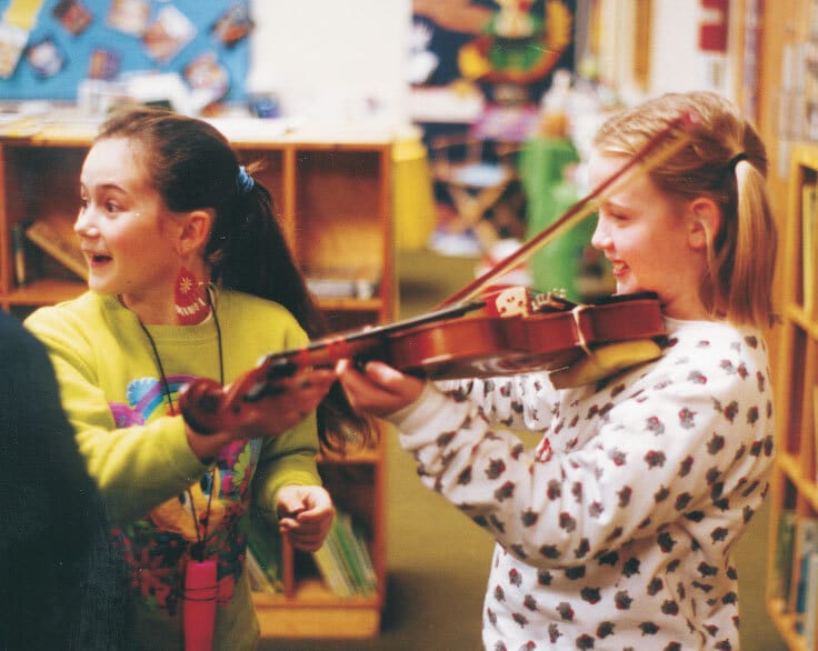 two girls standing in a classroom and smiling as one of the girls plays a violin.