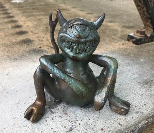 Metal statue of one-eyed mischievous creature sitting on pavement smiling